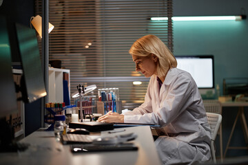 Middle aged Caucasian woman working at laboratory desk analyzing scientific samples under lamp...