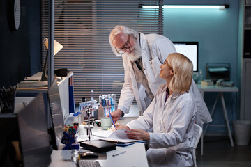 Senior Caucasian man standing and talking to middle aged Caucasian woman sitting at laboratory...