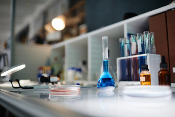 Laboratory table displaying glass beaker filled with blue liquid, petri dish containing pink substance, various test tubes and reagent bottles arranged in scientific research environment