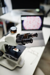 Vertical close up shot of laboratory microscope standing on desk with blurred laptop displaying...
