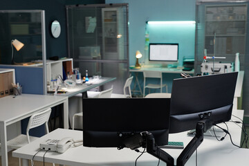 Empty scientific laboratory workspace showing modern computer monitors, telephone, laboratory equipment and organized desks with scientific instruments and documents in background