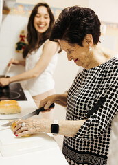 Grandmother slicing bread while granddaughter prepares hot chocolate in kitchen