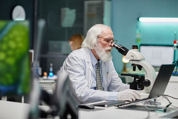 Senior Caucasian man with long hair and beard examining specimen through microscope in scientific...