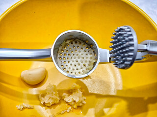 Garlic press resting on a yellow bowl with minced garlic and whole garlic clove nearby