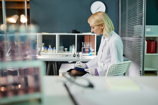 Middle aged Caucasian female scientist sitting at laboratory desk analyzing documents with laboratory glassware and scientific equipment in background during research process - Powered by Adobe