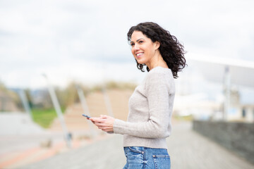 cheerful woman pauses during a walk, holding her phone and glancing back with a smile
