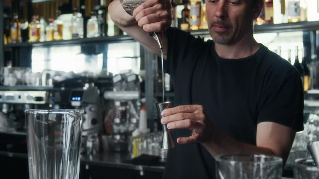 Medium shot of mature Caucasian man with tattoo pouring portion of vodka in jigger while making cocktail at bar counter