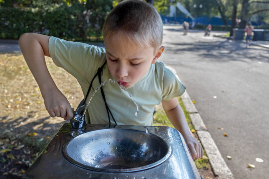 Young boy drinking from public water fountain on sunny hot day in park.