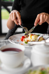 Close-up of a person cutting food on a white plate with vegetables and sauce, with a cup of tea on the table in a casual dining setting