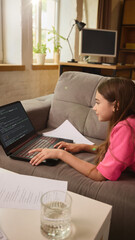 Teen girl lying on couch typing code on laptop, papers and glass of water on table, bright cozy...