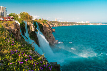  Scenic view of Duden Waterfalls in Antalya, Turkey, cascading from rocky cliffs into the Mediterranean Sea, with blooming purple flowers in the foreground and a turquoise sky.