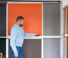 A man adjusts blinds with a remote in a modern showroom featuring multiple sun protection systems. The wall displays an array of styles, including pleated, roller, and venetian, highlighting their ver