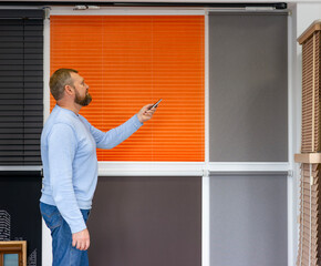 A man uses a remote control to operate motorized blinds in a showroom of window treatments store. He stands beside a display of pleated, roller, and wooden blinds in various colors and fabrics.