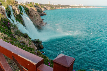  Scenic view of Duden Waterfalls in Antalya, Turkey, cascading from rocky cliffs into the Mediterranean Sea, with blooming purple flowers in the foreground and a turquoise sky.