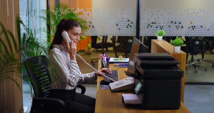 Indian young adult gen z woman lady sitting chair desk table use computer look screen do work task typing email check data at job office expert staff busy female girl attend phone calls talk day time