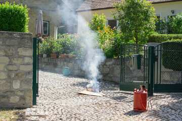 Small outdoor fire creating smoke in a paved backyard with houses and garden in the background,...