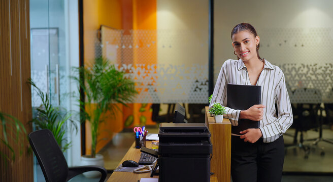 Indian happy young adult gen z proud woman lady standing hold file folder look camera do casual pose female girl worker expert staff staring cam at desk table indoor new job office work place