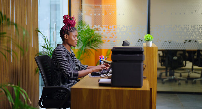 African young adult gen z woman lady sitting chair desk table use computer look screen do work task typing email check data at job office expert staff busy female girl attend customer day time