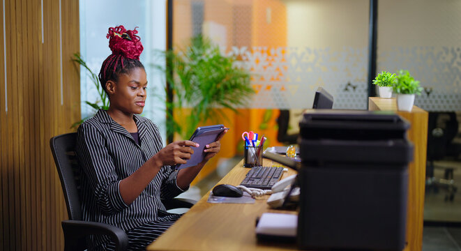 African young adult gen z busy woman lady sitting on chair use tablet device touch screen typing girl female agent hold gadget do work day task check email data read news play game job office place