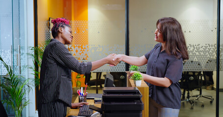 Happy young adult gen z woman lady staff agent standing at desk table do hand shake check in client visit indoor office smiling female girl talk share enquiry help  to visitor at job place day time