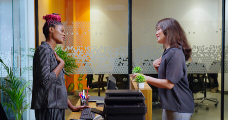 Happy young adult gen z woman lady staff agent standing at desk table do greeting check in client visit indoor office smiling female girl talk share enquiry help to visitor at job place day time