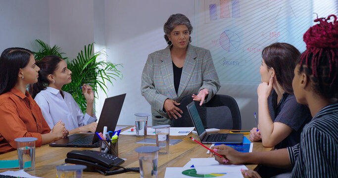 Indian old senior boss woman lady talk speak show white board chart learn study graph data growth work place. young adult female girl staff group team share stock trade new day idea plan job office