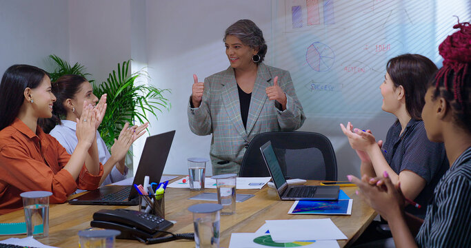 Indian old senior boss woman lady talk speak show thumb up white board chart learn study graph data work place. young adult girl staff group team share stock trade new day idea plan job office