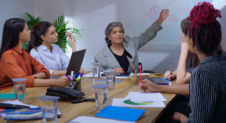 Indian young adult woman group sitting around table desk chair with boss CEO lady talk gossip enjoy work day indoor office female girl staff team group speak share good news spend break time job place