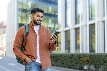 A smiling student, outdoors, using his phone while walking in front of a modern building