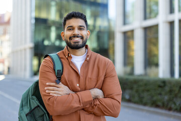 A smiling Indian student with a backpack stands confidently outside a modern building, with arms...