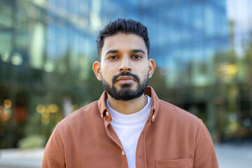 A portrait of a young man with a beard is taken in front of a modern building, creating a...
