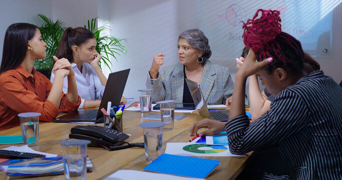 Indian young adult woman staff sitting around desk table with old aged boss ceo lady talk share bad news at job place upset female leader speak about work loss to girl group team office board room