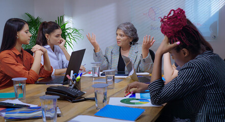 Indian young adult woman staff sitting around desk table with old aged boss ceo lady talk share bad news at job place upset female leader speak about work loss to girl group team office board room