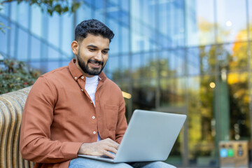 A smiling Indian man in an orange shirt works on a laptop outdoors, likely a freelancer or student in a modern urban setting.