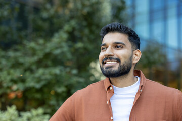 A smiling Indian man with a beard, dressed in a casual orange shirt, looks off-camera with green...