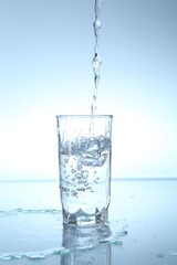 Pouring water into glass at mirror table against light blue gradient background, closeup