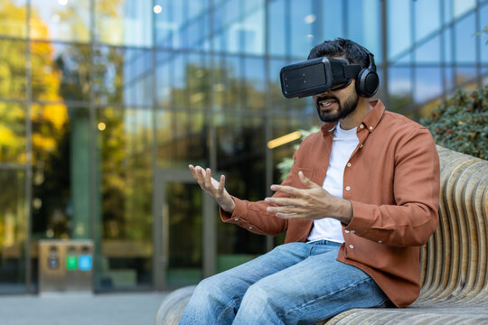 A man wearing a VR headset gestures excitedly outdoors, experiencing virtual reality in a modern setting with a building in the background.
