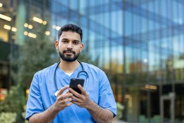 A doctor in scrubs holds a phone in front of a modern building, possibly using it for communication.