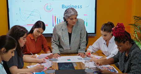 Indian old boss ceo woman lady show stock trade data on chart paper growth graph talk share idea plan speak work day task with young adult expert girl staff group intern team at job office room place	