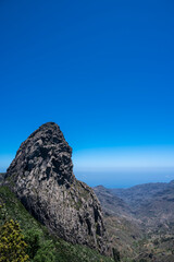 A mountain with a large rock on top and a blue sky in the background. The mountain is surrounded by trees and the sky is clear and bright