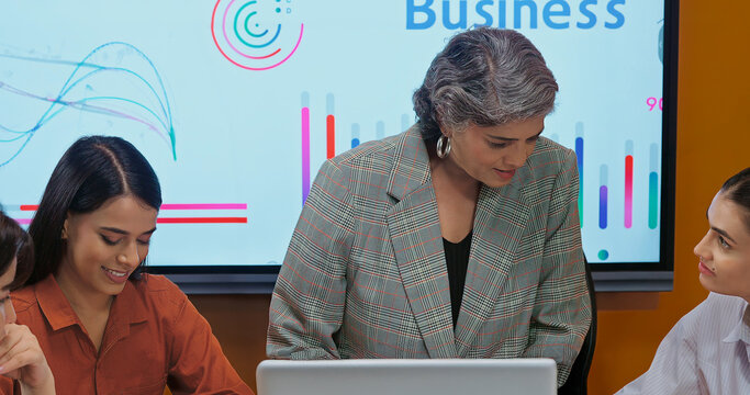 Indian asian young female old senior boss lady team leader show chart board learn study graph data growth work place adult girl talk speak share stock trade idea plan staff group job office day