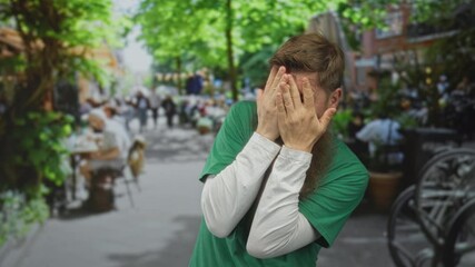 Bearded man in green volunteer shirt covers face with both hands on city street; anxiety frustration. - Powered by Adobe