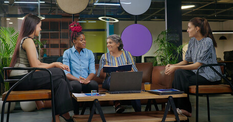 Indian old aged boss lady hold file do share good news new deal work talk speak ideas plan job place happy female girl staff group team sitting on chair sofa couch use laptop look screen at office