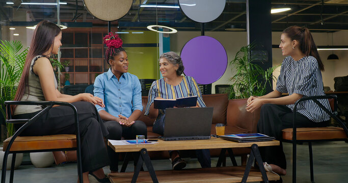 Indian old aged boss lady hold file do share good news new deal work talk speak ideas plan job place happy female girl staff group team sitting on chair sofa couch use laptop look screen at office - Powered by Adobe