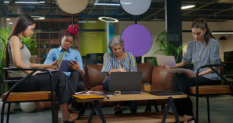 Indian busy old aged boss lady sitting on sofa couch use on desk table laptop look screen do work check email data job place young adult female staff expert girl team hold paper read report day time