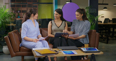 Indian happy busy woman leader young adult staff sitting sofa use tablet look screen check email talk work day task with team female girl expert hold paper listen share new ideas plan job office place