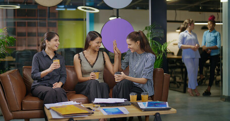 Indian asian smiling young adult gen z diverse girl woman sitting on sofa drink coffee indoor job office happy female intern staff talk funny gossip spend break time enjoy fun joy day at work place