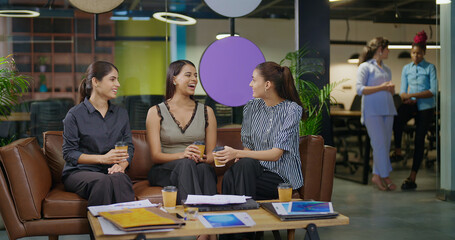 Indian young adult gen z woman lady group sitting on sofa hold cup drink coffee indoor job office happy female girl intern staff talk funny gossip spend break time enjoy fun joy day at job work place