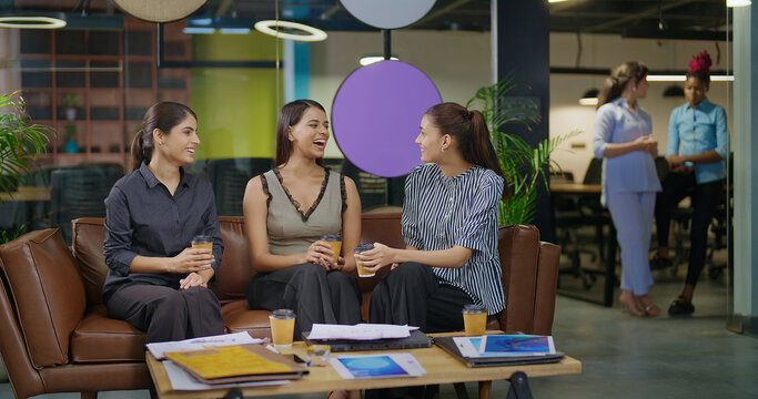 Indian young adult gen z woman lady group sitting on sofa hold cup drink coffee indoor job office happy female girl intern staff talk funny gossip spend break time enjoy fun joy day at job work place