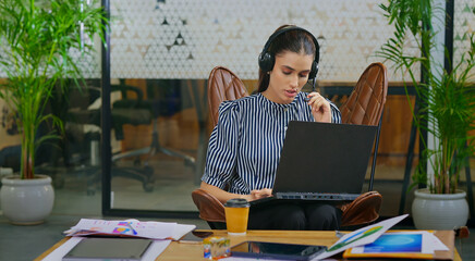 Indian young adult gen z busy woman lady sitting on chair wear headphone use laptop look screen do...